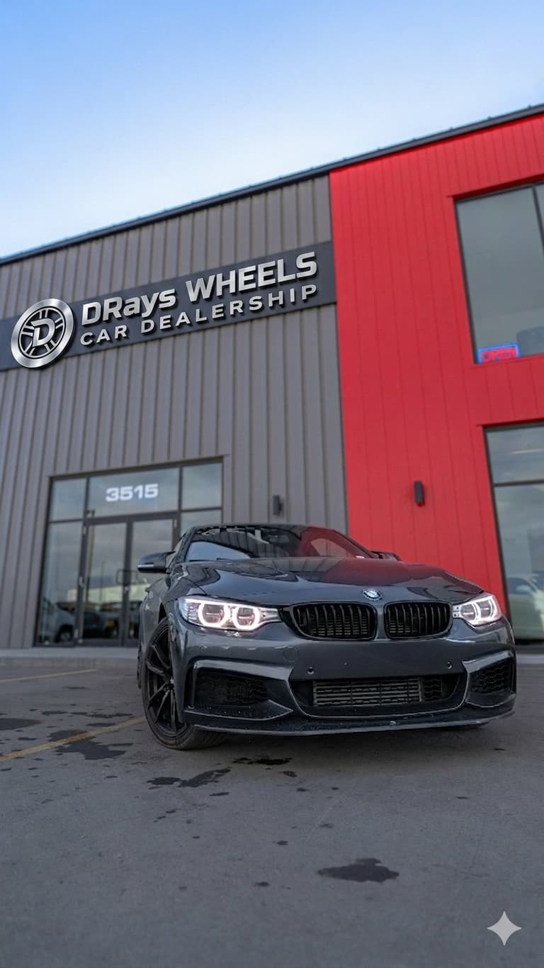 A sleek, glossy dark gray sports sedan parked prominently on the asphalt in front of the modern Drays Wheels Car Dealership building facade. The building features gray corrugated siding, a bright red accent section, and a large metallic silver brand sign.